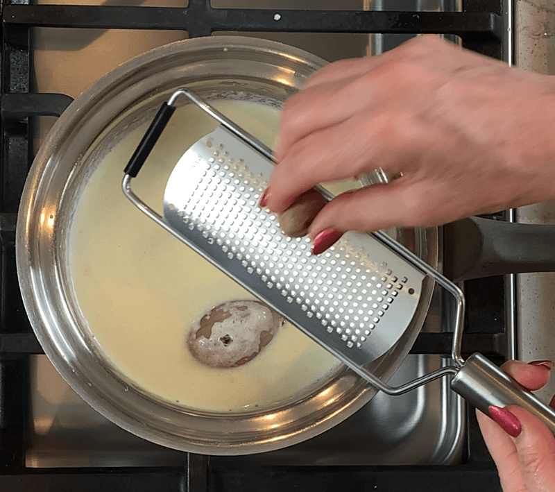Fresh nutmeg being grated with a microplane directly over a simmering cream sauce
