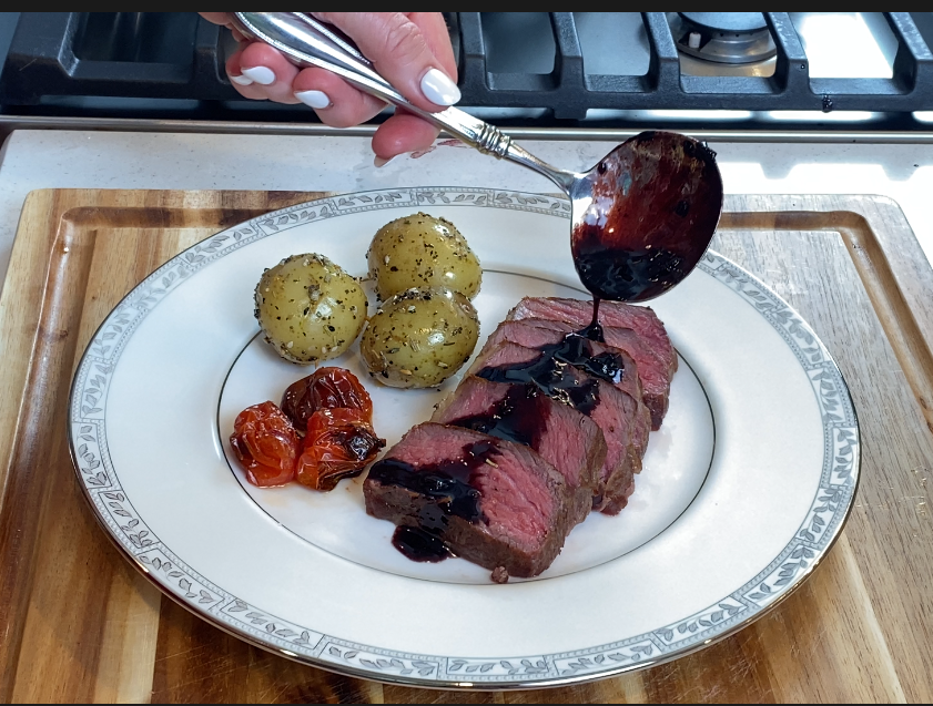 Sliced steak on a white plate with red wine shallot reduction sauce poured over the top, served with roasted potatoes and tomatoes