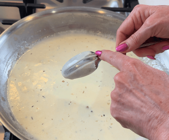 A finger drawing a clean line through cream sauce on the back of a spoon to test if reduced enough