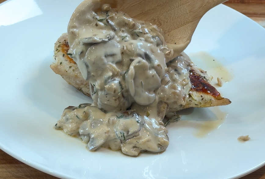 Pouring rich mushroom cream sauce from a wooden spoon over pan-seared chicken on a white plate