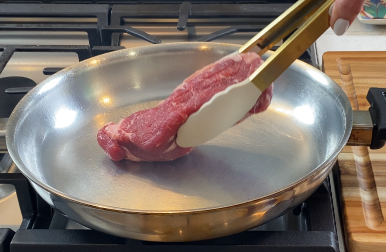 Placing a raw NY strip steak into a hot stainless steel skillet to sear