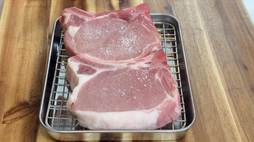 Two seasoned bone-in pork chops resting on a wire rack before searing