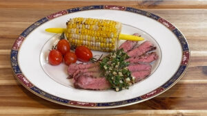 Chimichurri-inspired butter sauce poured over sliced flank steak with grilled corn on the cob and roasted tomatoes.