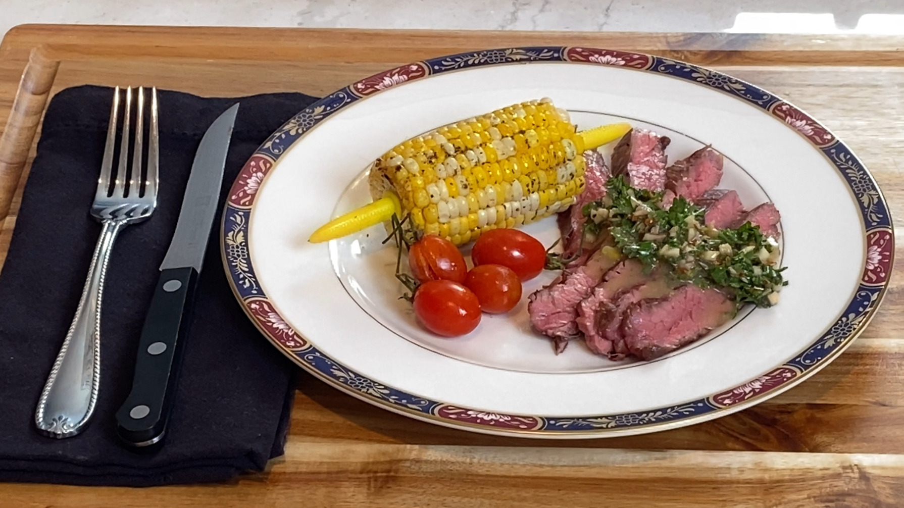 Sliced flank steak with chimichurri-inspired butter sauce on top, paired with corn on the cob and roasted tomatoes.