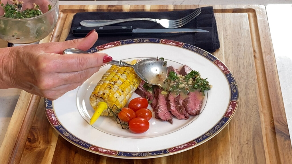 Fanned flank steak slices being topped with chimichurri-inspired butter sauce paired with grilled corn on the cob and roasted red tomatoes.