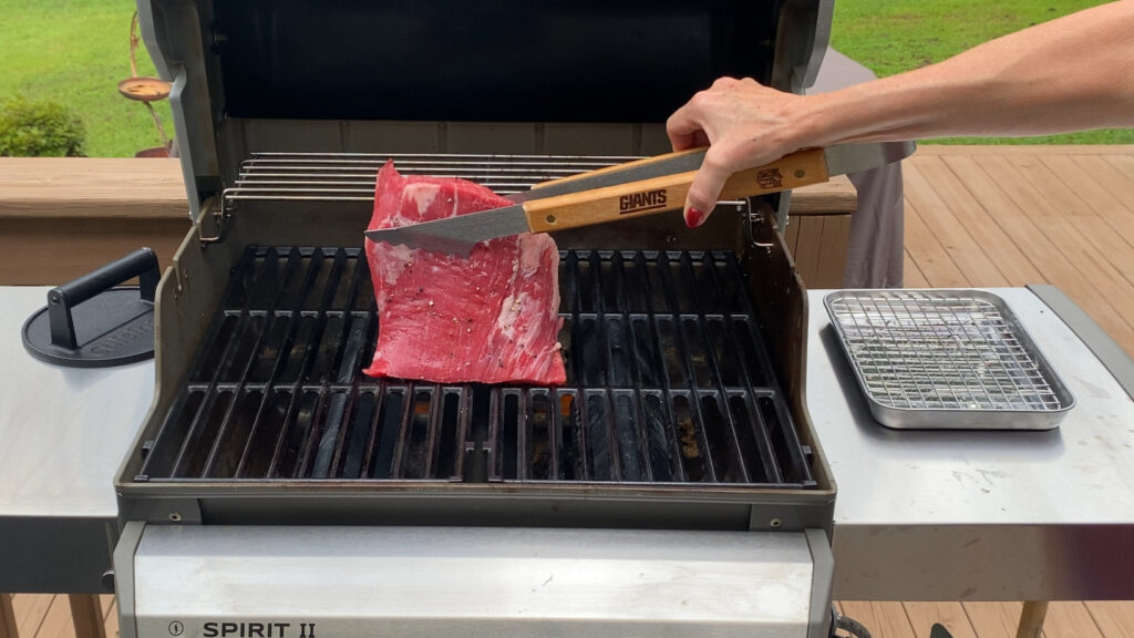 Raw flank steak seasoned with salt and pepper being placed on a hot grill