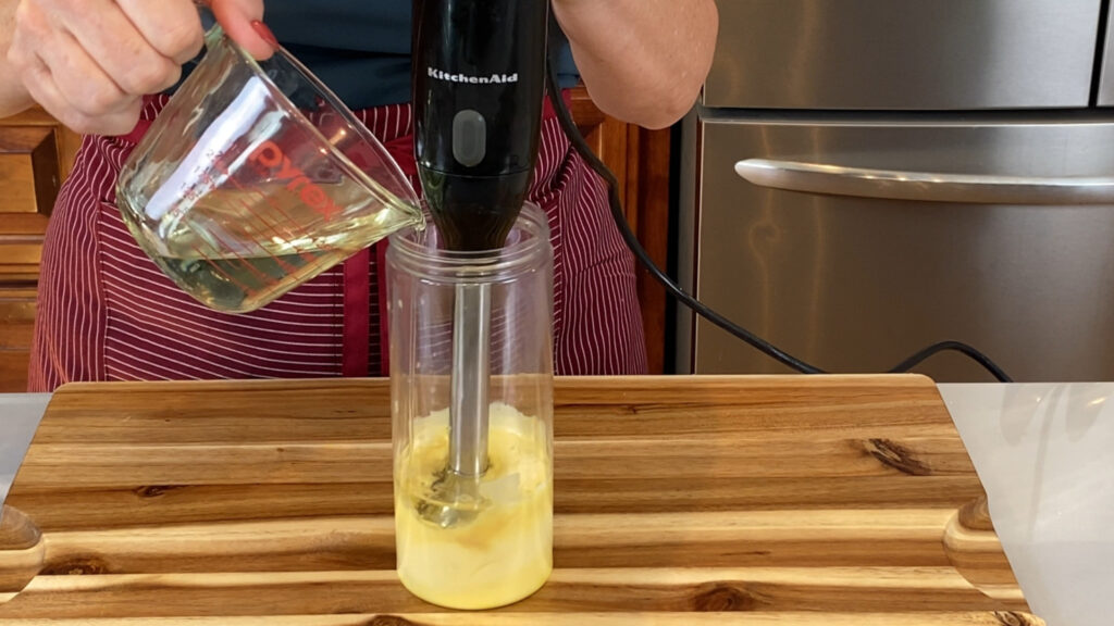 Oil being poured on top of the yolk mixture with an immersion blender in a narrow container.