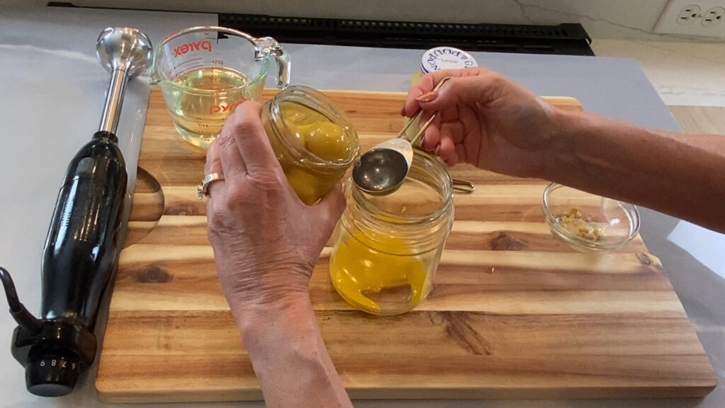 Preserved lemon brine being added to an egg yolk and mustard mixture with oil and an immersion blender ready to make it into homemade mayonnaise. 