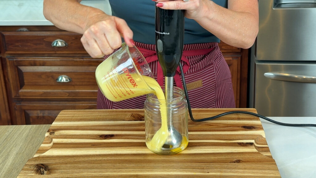 Broken egg emulsion mixture being added to a fresh yolk to re-emulsify using an immersion blender.